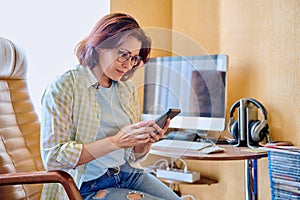 Middle aged woman using smartphone sitting in armchair in home office