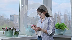 Middle aged woman using smartphone, standing near panoramic window