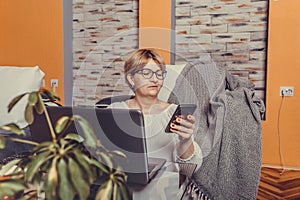 Middle aged woman using smartphone and working on laptop at home