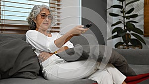 Middle aged woman watching tv on comfortable sofa, enjoying carefree leisure weekend activity at home