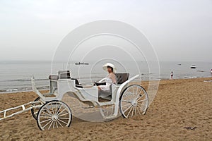 Middle-aged woman in a white dress and a hat in a white open carriage on a background of the sea coast