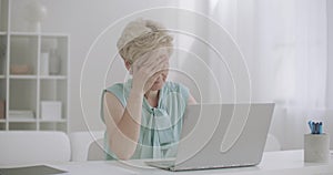 Middle-aged woman is working with laptop is feeling headache, touching forehead by hands, overexertion and fatigue