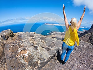 Middle-aged woman in yellow t-shirt and jeans raising her arms to the sky on top of a cliff on a sunny day