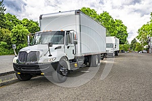 Middle class rig semi trucks with box trailers standing on the city street in industrial area