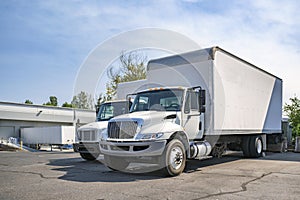 Middle duty compact rig semi trucks with box trailers standing on the warehouse parking lot take a break between freights