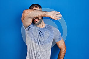 Middle east man with beard standing over blue background covering eyes with arm, looking serious and sad