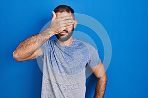 Middle east man with beard standing over blue background covering eyes with hand, looking serious and sad