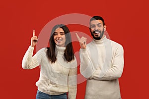 Middle eastern couple pointing fingers up, having great idea and smiling to camera, standing over red studio background