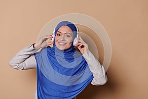 Middle-Eastern Muslim woman in blue hijab, listens to music on headphones, smiles at camera, isolated beige background