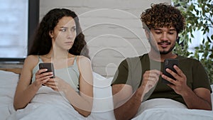 Middle eastern wife spying her cheating husband texting on cellphone, having flirty chat conversation with lover in bed
