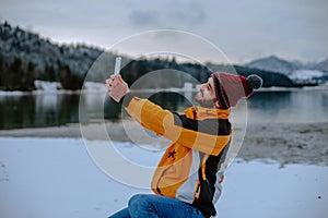 In the middle of nature with a big lake and snowy mountain and forest young man with orange jacket taking pictures of