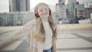 Middle shot of cheerful young woman in headphones dancing on urban city square on cloudy autumn or spring day. Portrait