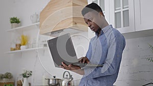 Middle shot of concentrated young man standing in kitchen typing on laptop keyboard. Portrait of focused African