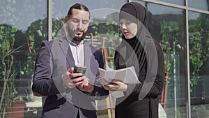 Middle shot of confident muslim woman with documents and man with smartphone discussing business deal outdoors. Portrait