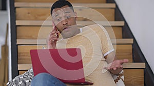 Middle shot front view of thoughtful African American man talking on the phone typing on laptop keyboard sitting on