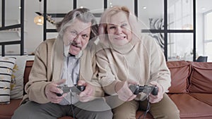 Middle shot of funny senior couple playing video games. Portrait of cheerful Caucasian husband and wife using game