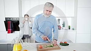 Middle shot of handsome senior man slicing cucumber for healthful salad as blurred cheerful woman talking on the phone