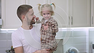 Middle shot of happy Caucasian father kissing baby daughter looking at camera and smiling. Portrait of cheerful handsome