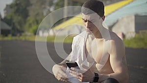 Middle shot of perspiring sportsman with smartphone wiping face with white towel. Portrait of fit handsome Caucasian man