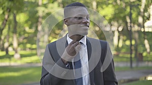 Middle shot portrait of confident businessman in suit adjusting necktie and smiling at camera. Young African American