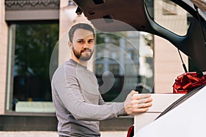 Middle shot portrait of delivery man standing near car, large box with beautiful red bow at truck.