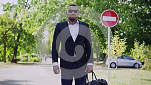 Middle shot portrait of handsome elegant African American man in formal suit and sunglasses standing outdoors with bag