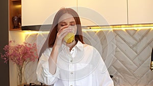 Middle shot portrait of happy young woman in casual clothes drinking orange juice from glass standing in modern kitchen