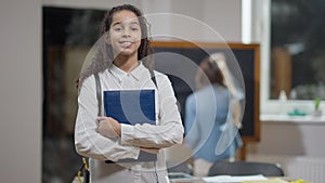 Middle shot portrait of positive cute African American schoolgirl with workbook posing in classroom with blurred