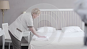 Middle shot of serious maid putting clean towels on white bed and leaving. Side view portrait of professional Caucasian