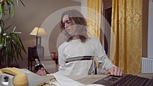 Middle shot of skinny 1990s young man using vintage laptop and training biceps sitting at table indoors. Portrait of