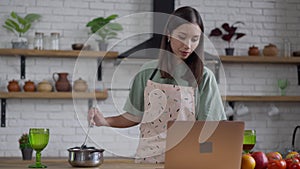 Middle shot of smiling young slim Asian woman mixing sauce in cooking pan looking at laptop screen. Portrait of