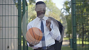 Middle shot of young African American businessman standing at the entrance to outdoor basketball court and looking at