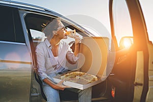 Midle-aged woman eating just cooked italian pizza sitting on driver car seat during meal break and enjoying sunset light. Auto