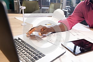 Midsection of african american mature engineer using laptop while sitting with blueprint at desk