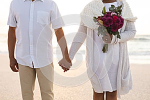 Midsection of biracial man holding hands with woman holding flower bouquet at beach wedding ceremony