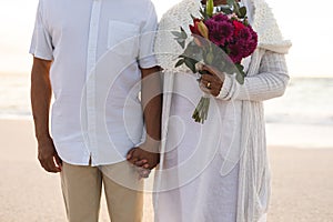 Midsection of biracial senior woman holding flower bouquet and man's hand at beach wedding ceremony