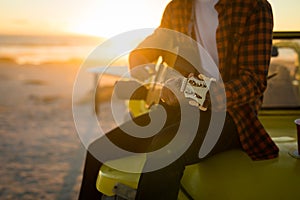 Midsection of caucasian man sitting on beach buggy by the sea playing guitar during sunset