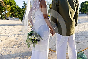 Midsection of caucasian newlywed couple holding hands and standing at beach at wedding ceremony