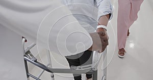 Midsection of diverse male doctor helping senior female patient use walking frame, slow motion