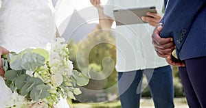 Midsection of diverse man with tablet officiating marriage of couple in sunny garden slow motion