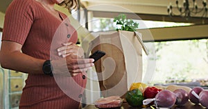 Midsection of happy caucasian pregnant woman touching belly, using smartphone in kitchen