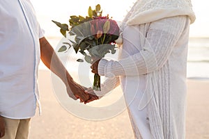 Midsection of multiracial senior couple holding hands with bouquet during wedding ceremony at beach