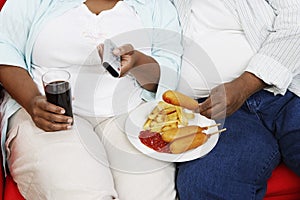 Midsection Of Overweight Couple With Junk Food Holding Remote Control