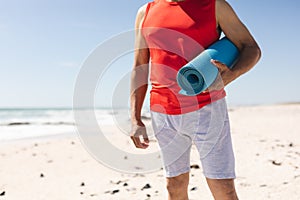 Midsection of retired senior biracial man holding rolled yoga mat at beach on sunny day