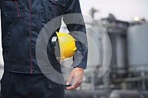 Midsection of young worker holding a yellow hardhat outdoors with factory in the background