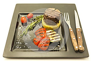 Mignon steak with fried potatoes and grilled vegetables on a beautiful black plate with knife and fork on a white background.