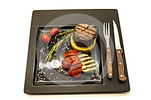 Mignon steak with fried potatoes and grilled vegetables on a beautiful black plate with knife and fork on a white background.