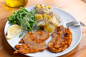 Milanesa steak or breaded schnitzel served on dish with green salad and cooked potato salad on wooden table. homemade simple lunch