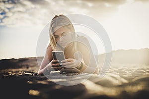 Milennial young beautiful blonde girl type on the phone lay down on the sand at the beach in outdoor nature activty - technology