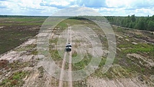 A military camouflaged passenger car is driving along a dirt road among forests and fields, aerial view from above. Concept: speci